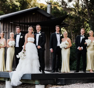 A group of people in formal attire, including a bride in a white gown and groom in a tuxedo, standing on a wooden platform in front of a small building with trees in the background.