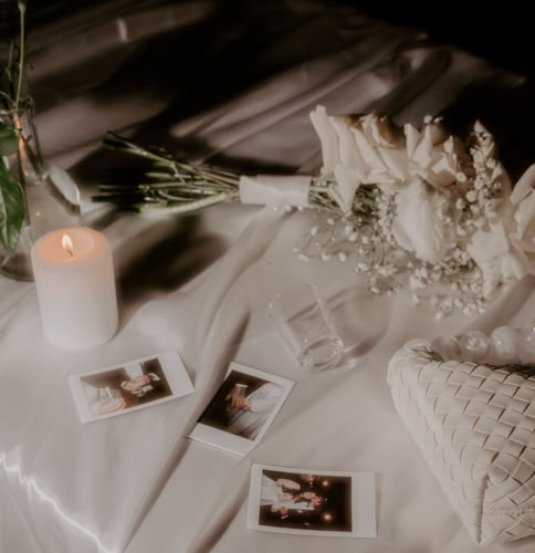 A lit candle, white flowers, and several instant photos on a white cloth surface.