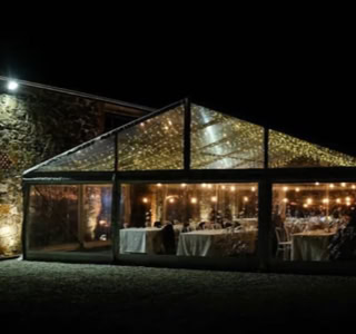A transparent tent with string lights inside, set up outdoors at night, with tables and chairs visible through the tent walls.